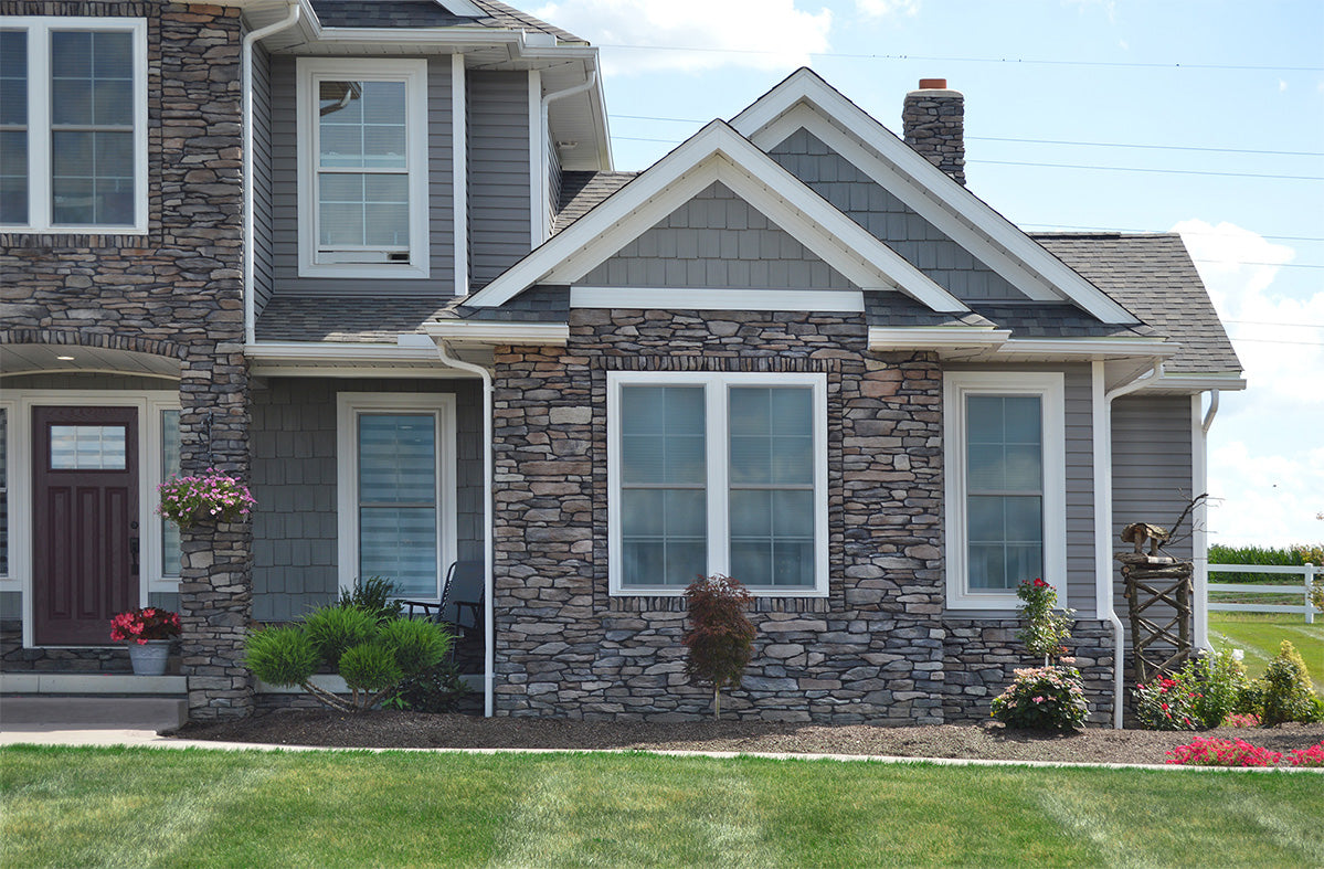 Two-story house with stone facade and gray siding, surrounded by green grass and flowers.