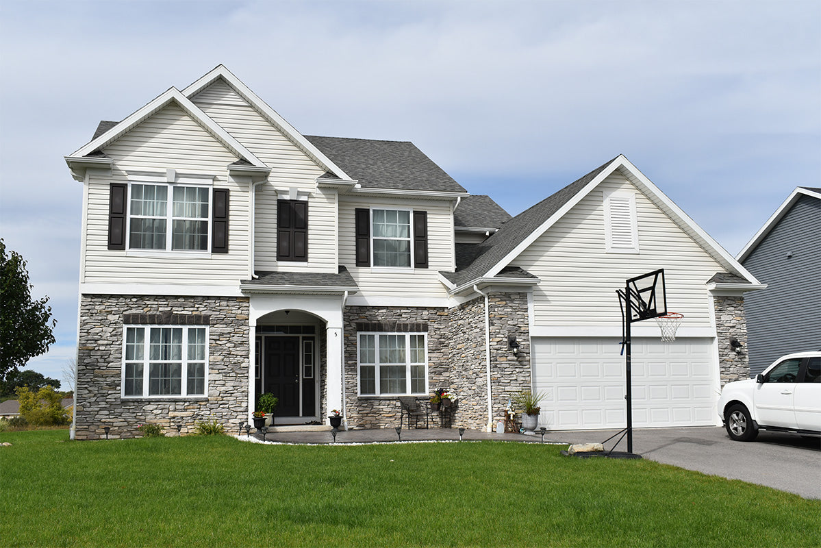 Two-story house with stone facade and white siding, featuring a basketball hoop on a clear day.