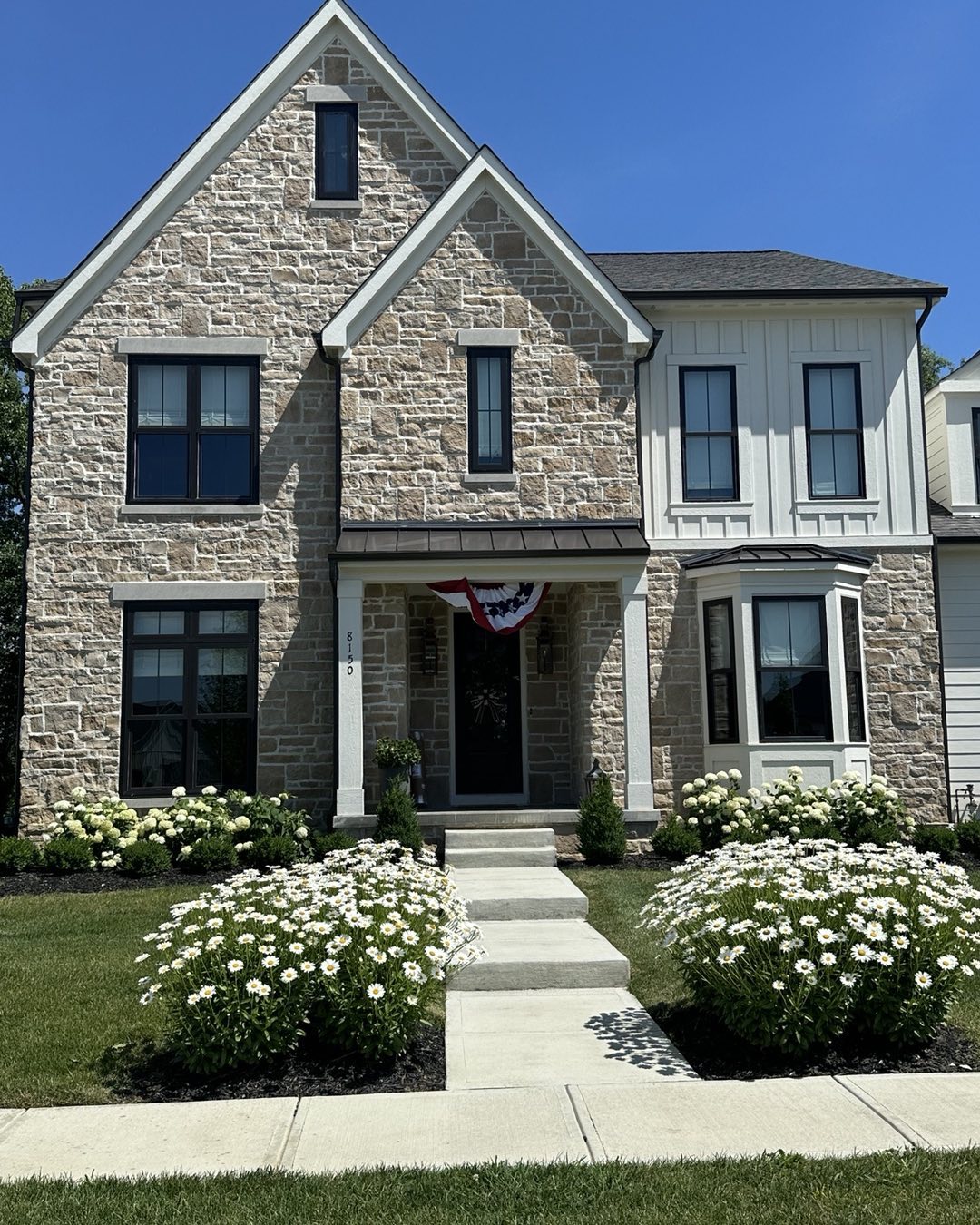 Two-story stone house with a front porch and garden beds filled with white flowers.