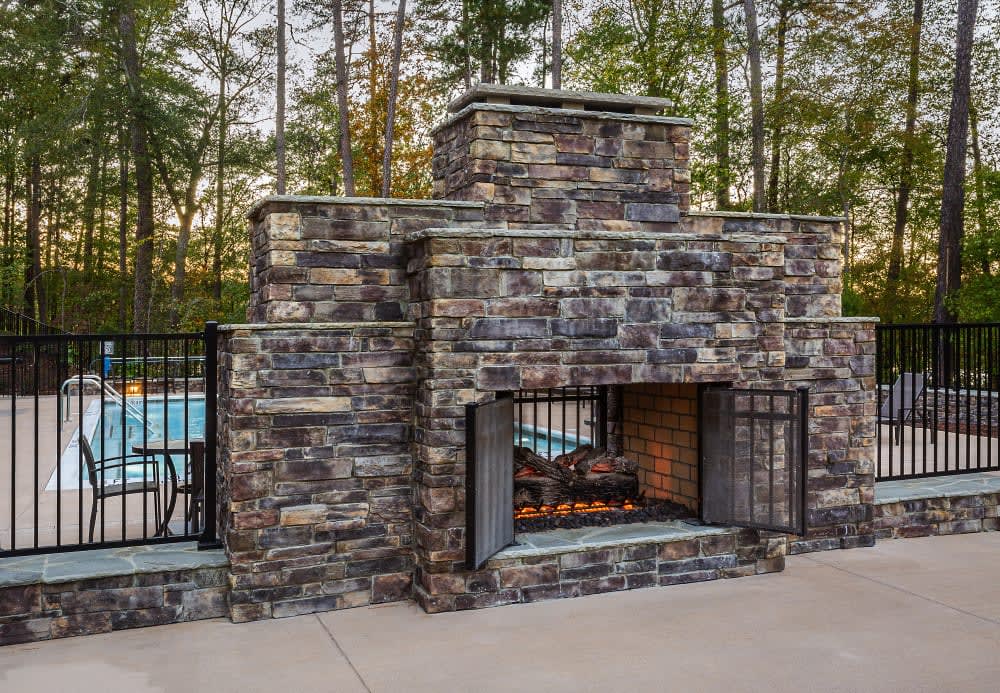 Ledgestone outdoor fireplace on a paved terrace beside a pool fence and wooded backdrop, featuring Cultured Stone Wolf Creek Country Ledgestone.