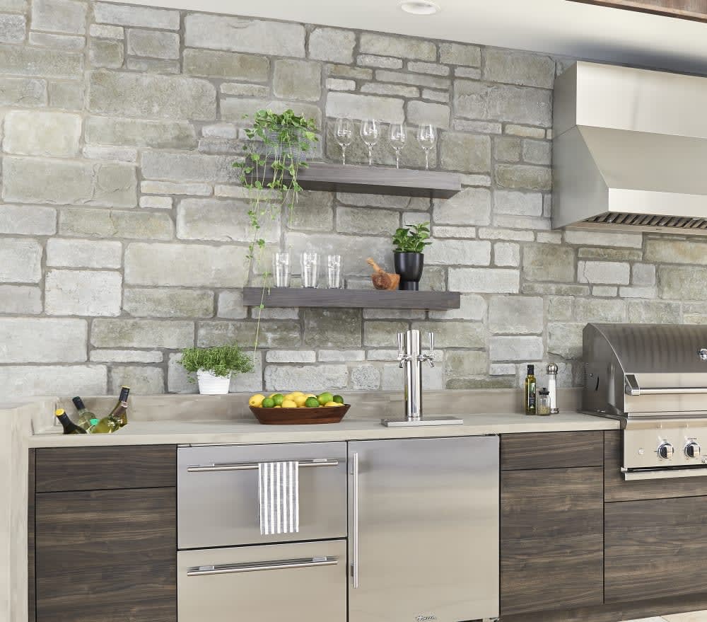 Light ashlar stone backsplash and wall behind a kitchen sink, cabinets and range, featuring Cultured Stone Silver Shore Sculpted Ashlar.