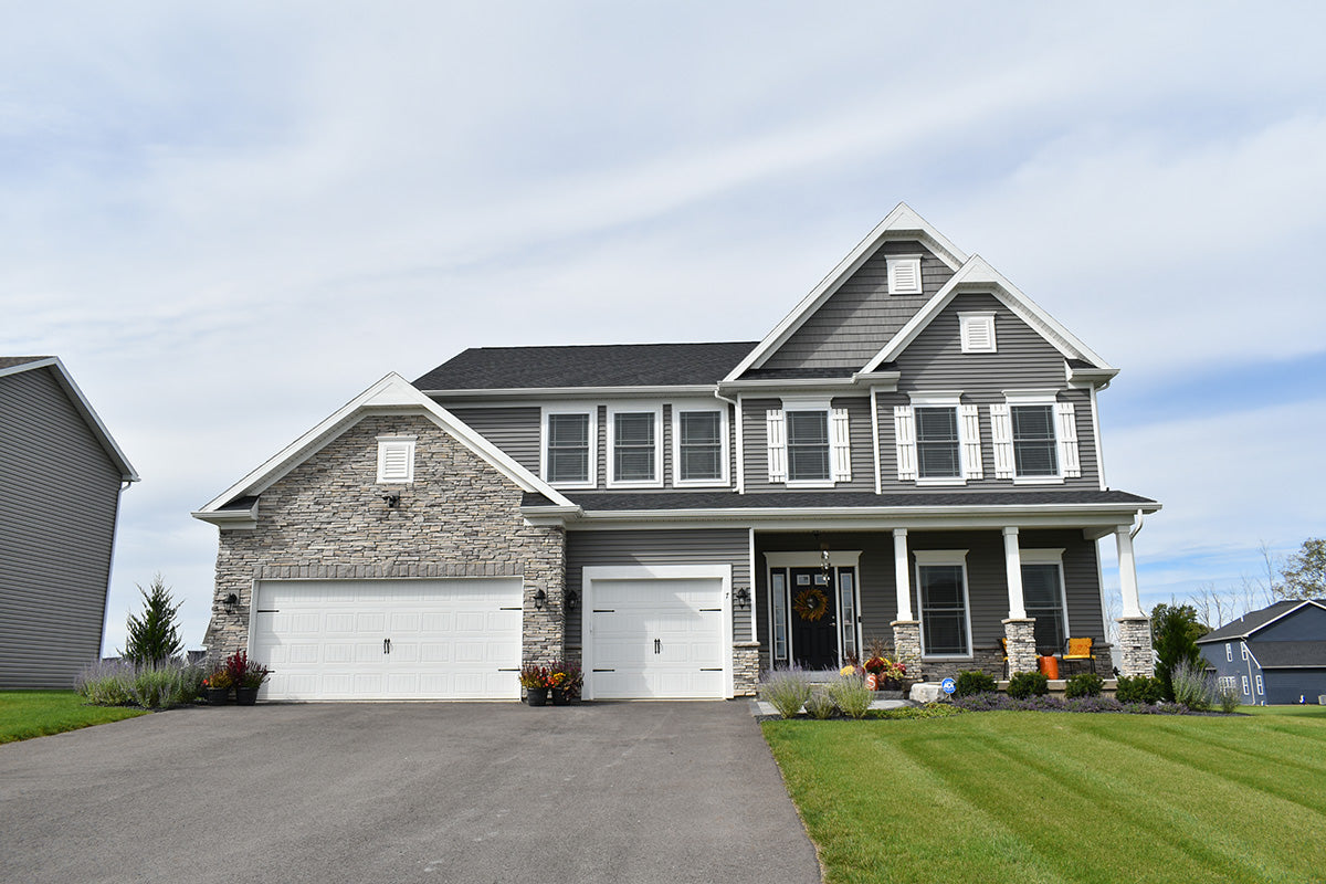 Two-story house with gray siding and a white garage door, surrounded by green grass and trees.