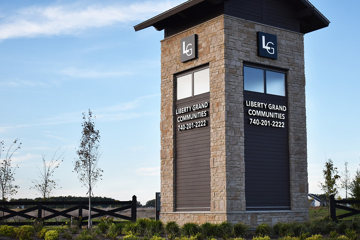 Stone tower with Liberty Grand Communities branding and contact information against a blue sky.
