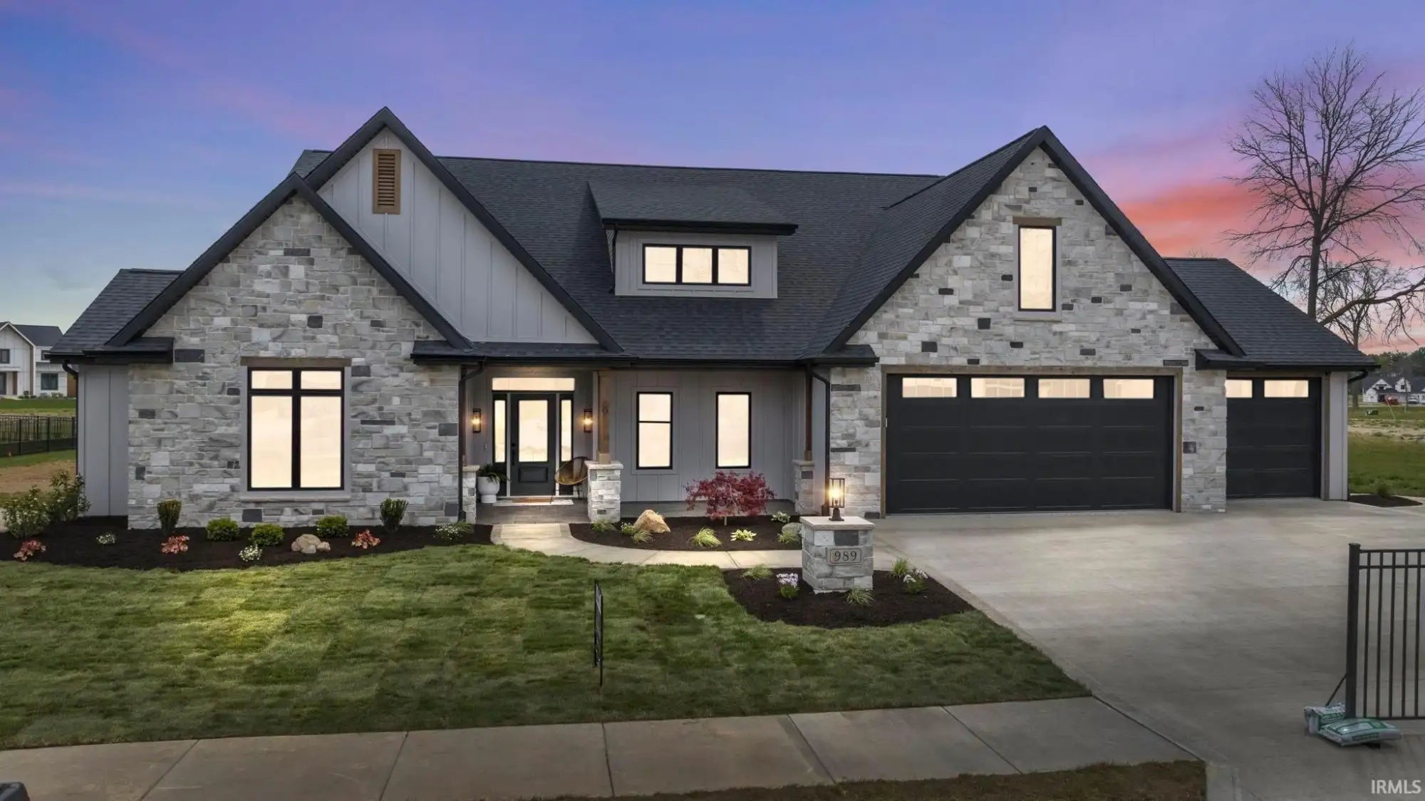 Modern gray stone house with black accents and garage doors, illuminated at dusk.